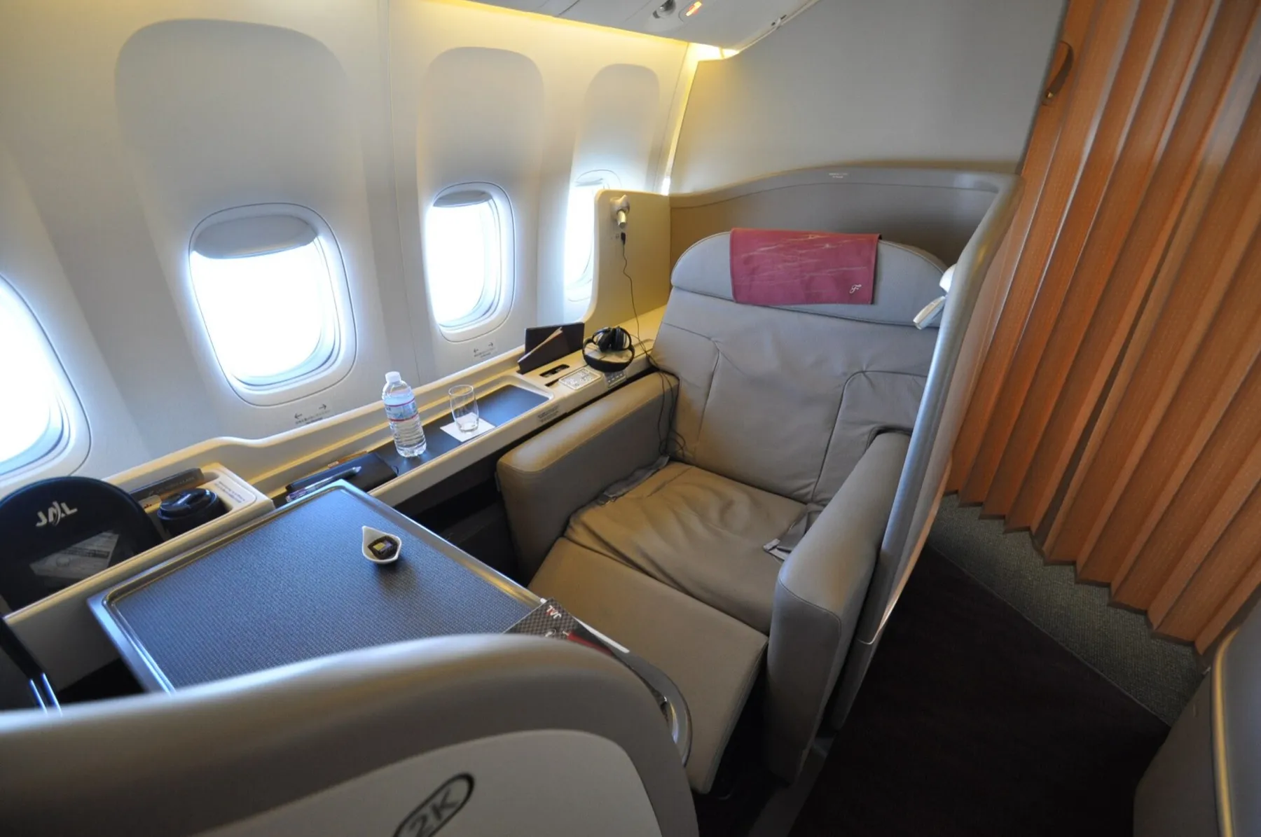 A JAL First Class suite by the window: a wide leather seat with a footrest, a dining table and a privacy divider