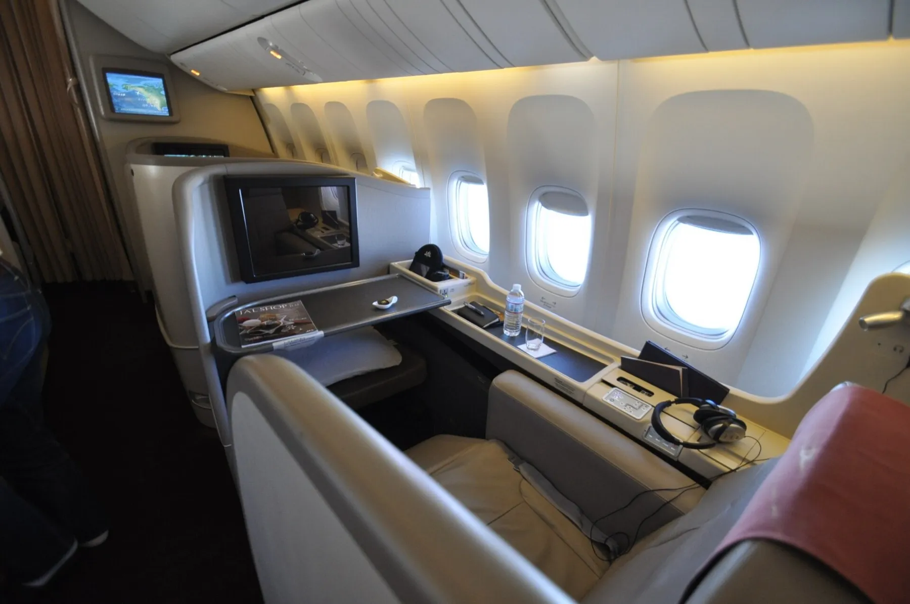Looking across a JAL First Class suite toward the windows, with a large personal screen, headphones and a dining table